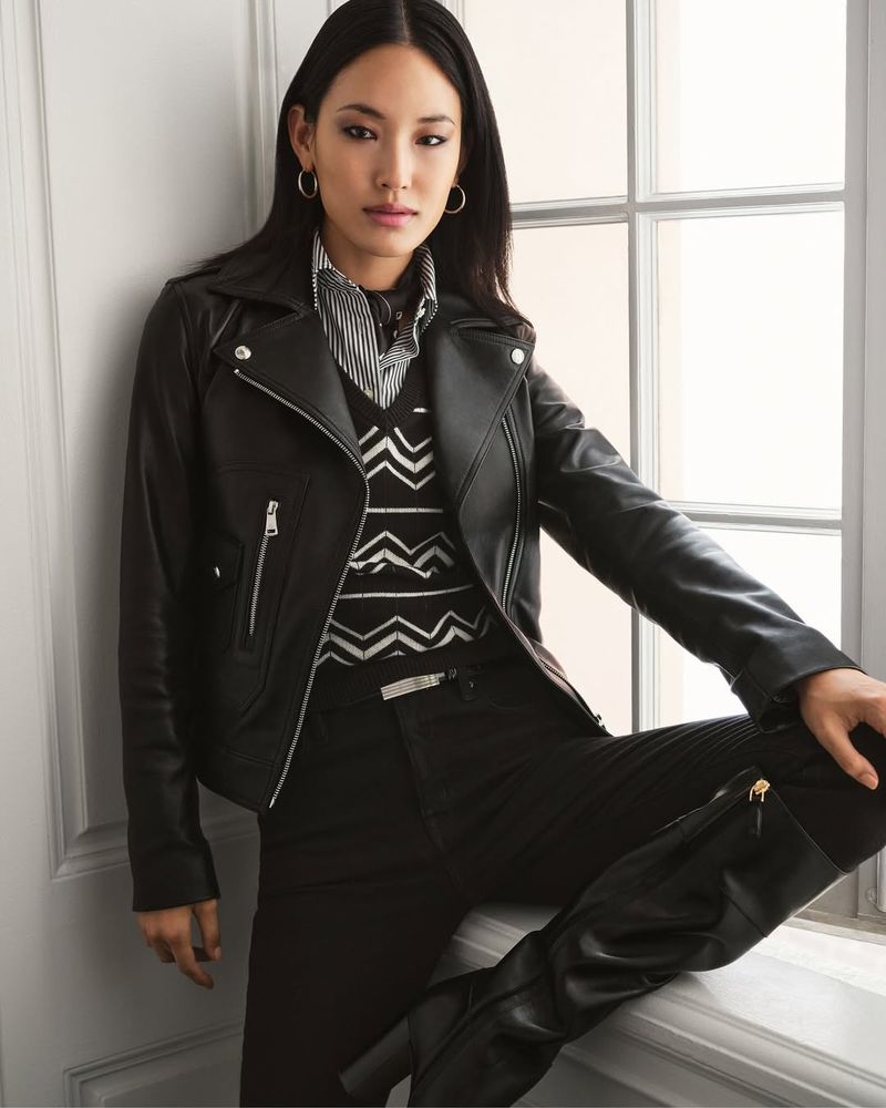 An Asian woman poses in a black leather jacket with black pants and knee-high boots, sitting by a bright window with a white frame.