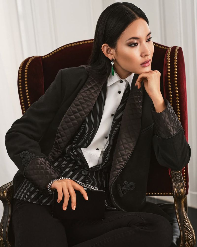 A woman in a black jacket and striped vest sits in a burgundy chair with a black clutch, looking to the side in a fashion-style studio shot.
