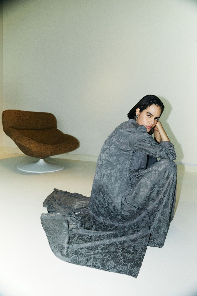 Model in patterned grey dress crouching on white floor with brown armchair in background