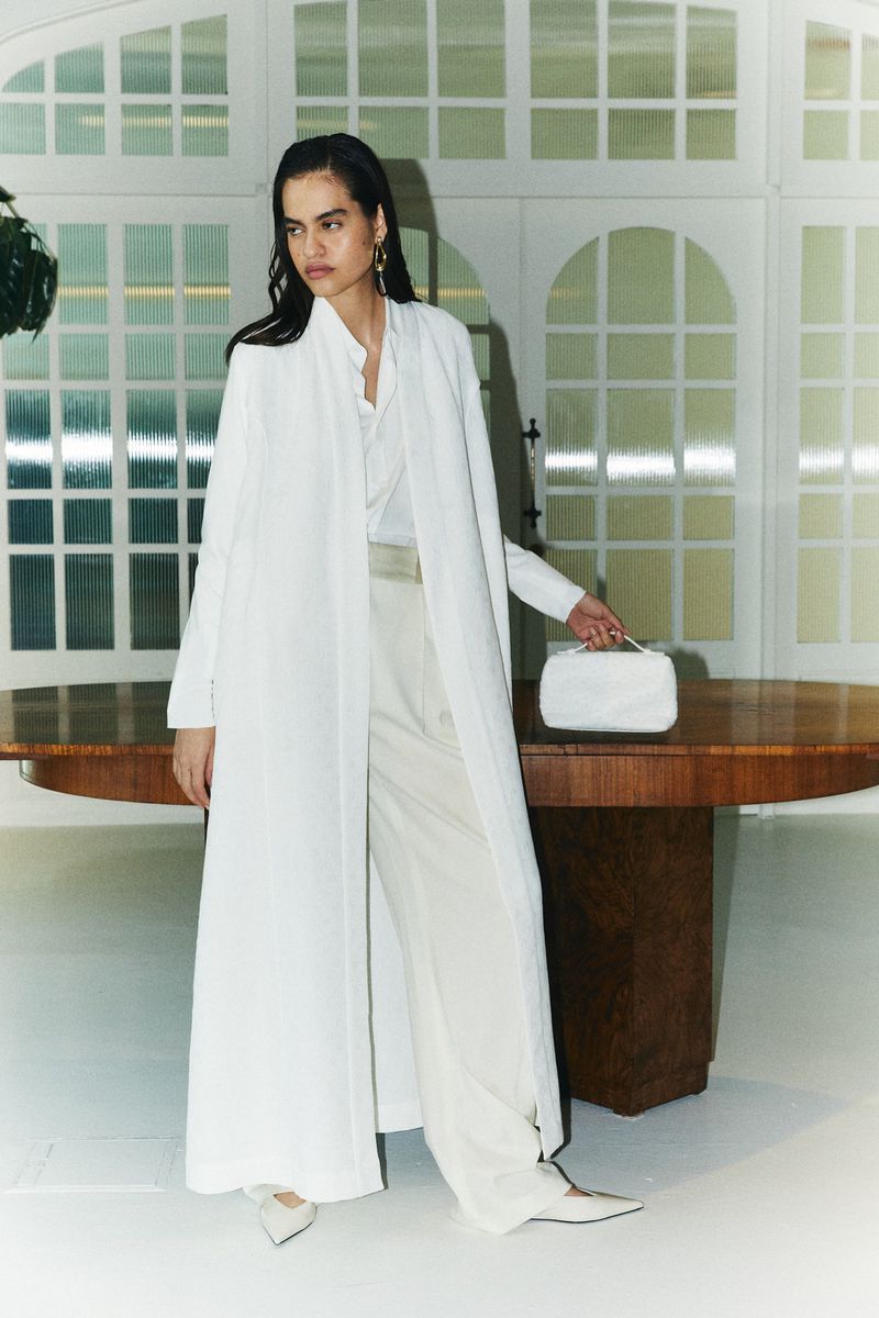 Elegant woman in long white coat and matching pants holds a white handbag, posing indoors with natural light and a neutral background.