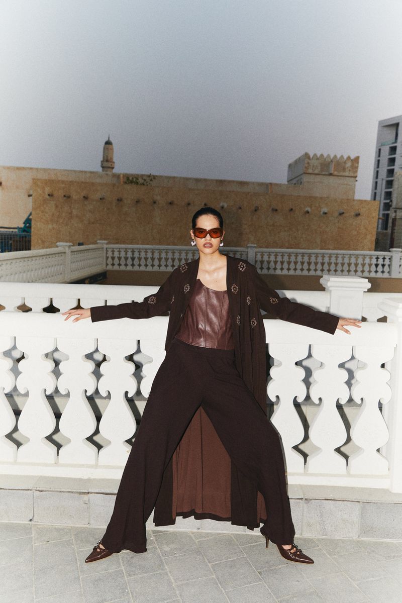 Fashionable adult female posing outdoors on a rooftop wearing sunglasses, a brown top, wide leg pants, and a long jacket, on a sunny day.