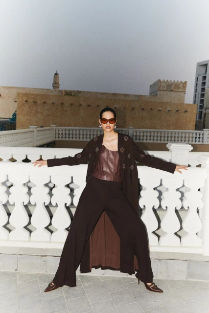 Fashionable adult female posing outdoors on a rooftop wearing sunglasses, a brown top, wide leg pants, and a long jacket, on a sunny day.