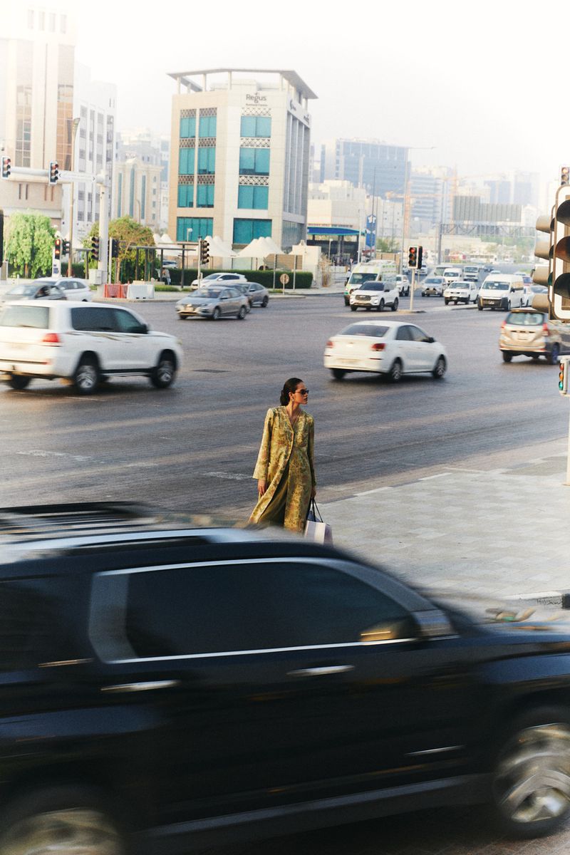 Adult female wearing a dress crosses a busy city street with cars and buildings in the background on a bright, sunny day.