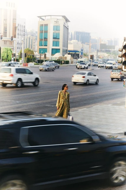 Adult female wearing a dress crosses a busy city street with cars and buildings in the background on a bright, sunny day.