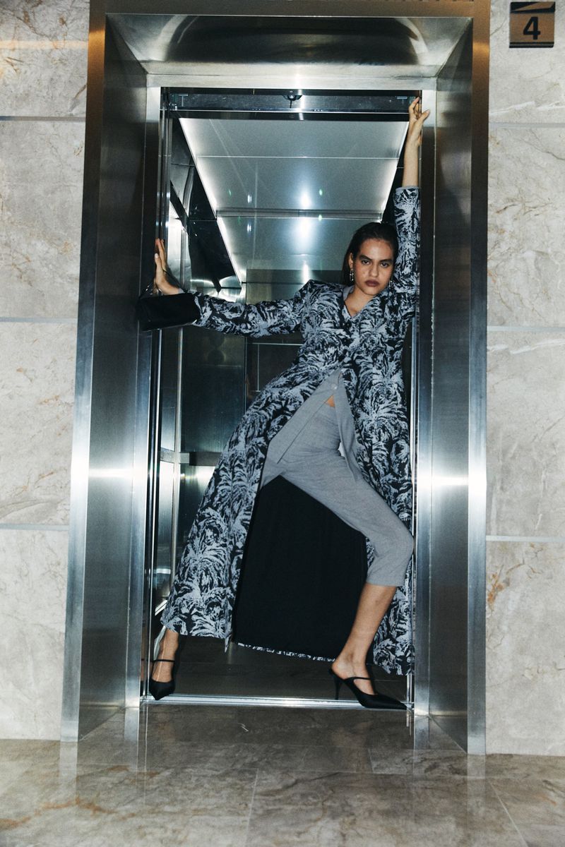 A tall fashion model poses in an elevator wearing a printed coat, grey sweatpants, and black heels, striking a pose with raised arms.
