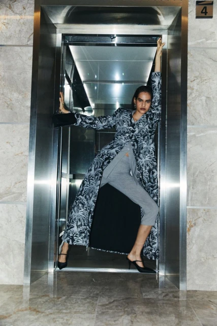 A tall fashion model poses in an elevator wearing a printed coat, grey sweatpants, and black heels, striking a pose with raised arms.