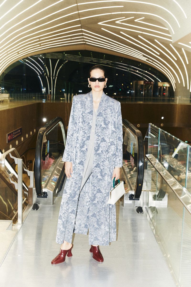 A woman in a patterned coat and sunglasses stands on an escalator in an airport or train station. Modern interior with bright lighting.