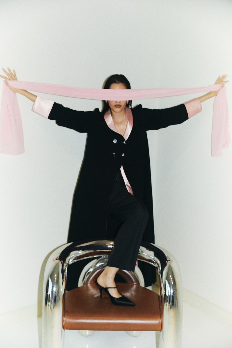 A woman in a black robe and pants sits on a chrome chair holding a pink fabric strip in front of her eyes in a studio shot.