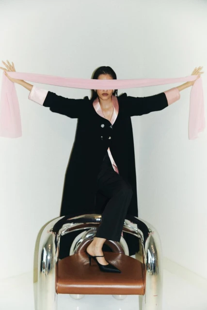 A woman in a black robe and pants sits on a chrome chair holding a pink fabric strip in front of her eyes in a studio shot.