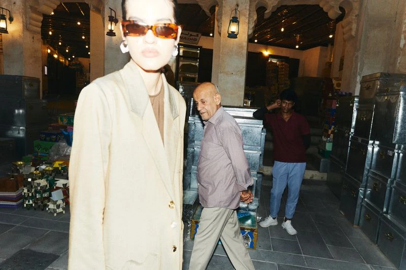 Fashionable woman in sunglasses and blazer stands out in an indoor market, complemented by two men and metallic storage containers.