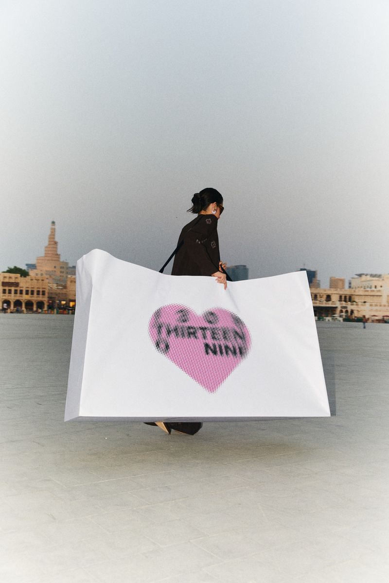 Woman holds large white board with pink heart over waterfront cityscape with historic buildings under cloudy skies in Doha, Qatar.