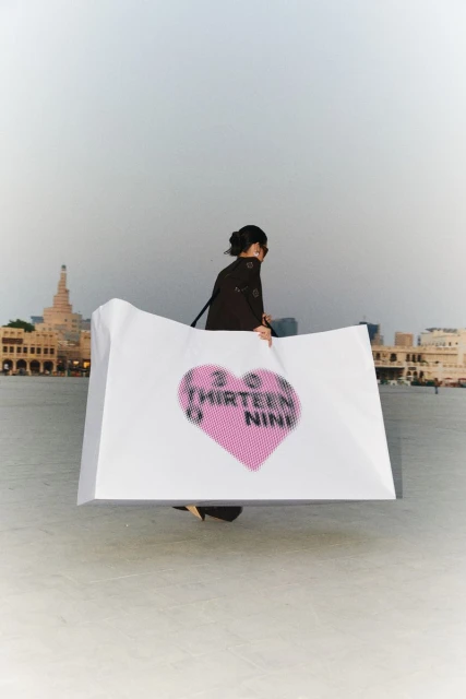 Woman holds large white board with pink heart over waterfront cityscape with historic buildings under cloudy skies in Doha, Qatar.