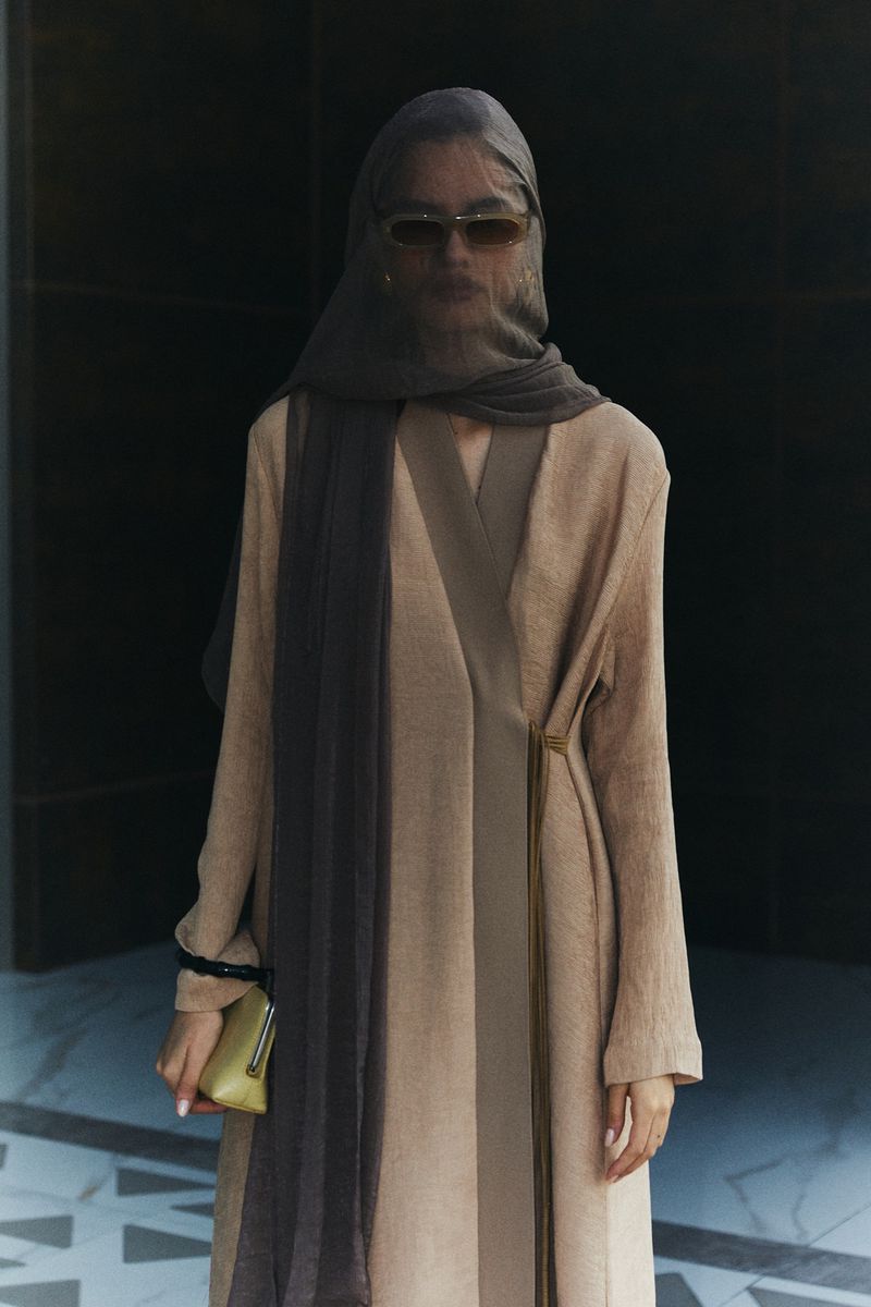 Adult female wearing a headscarf and long dress poses in front of a black wall, holding a small gold purse, in a fashion editorial style.