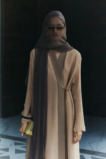 Adult female wearing a headscarf and long dress poses in front of a black wall, holding a small gold purse, in a fashion editorial style.