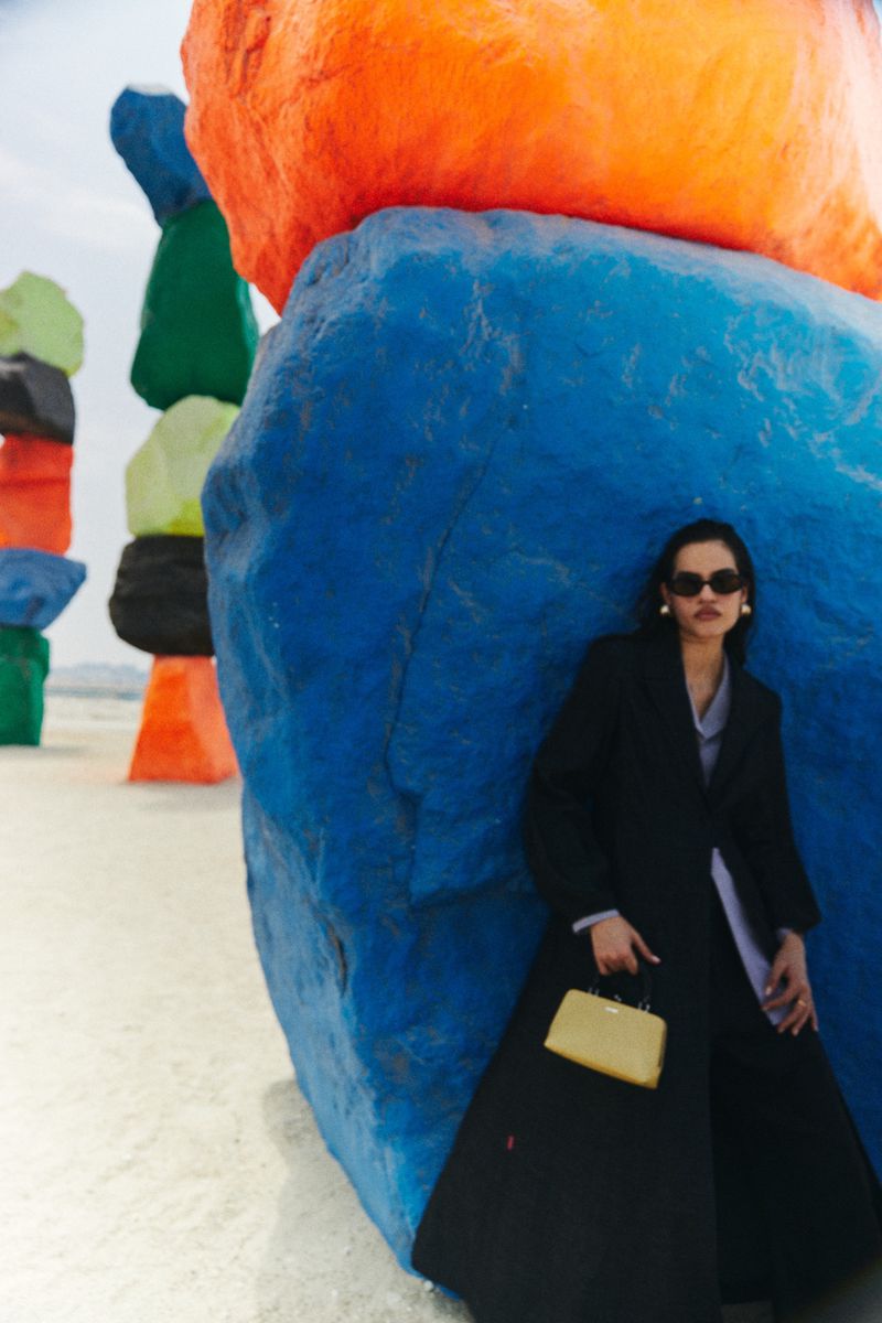Adult female in a black coat poses next to large, colorful rock formations in an outdoor setting with a tan purse.