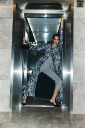 Model wearing patterned long coat and grey trousers posing inside an elevator doorway