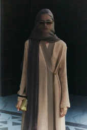 Model wearing a tan coat and sheer grey headscarf holding a small clutch bag