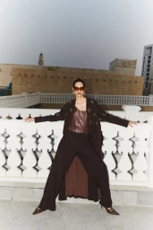 Model in brown coat and sunglasses posing on a rooftop with white balustrade and traditional architecture