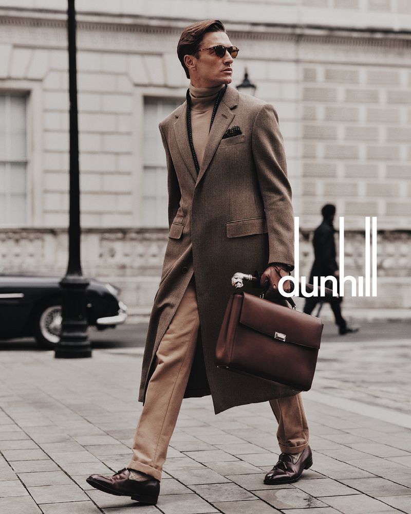 A man in a brown coat walks down a city sidewalk carrying a leather briefcase in a fashion editorial-style image with a vintage car in the background.