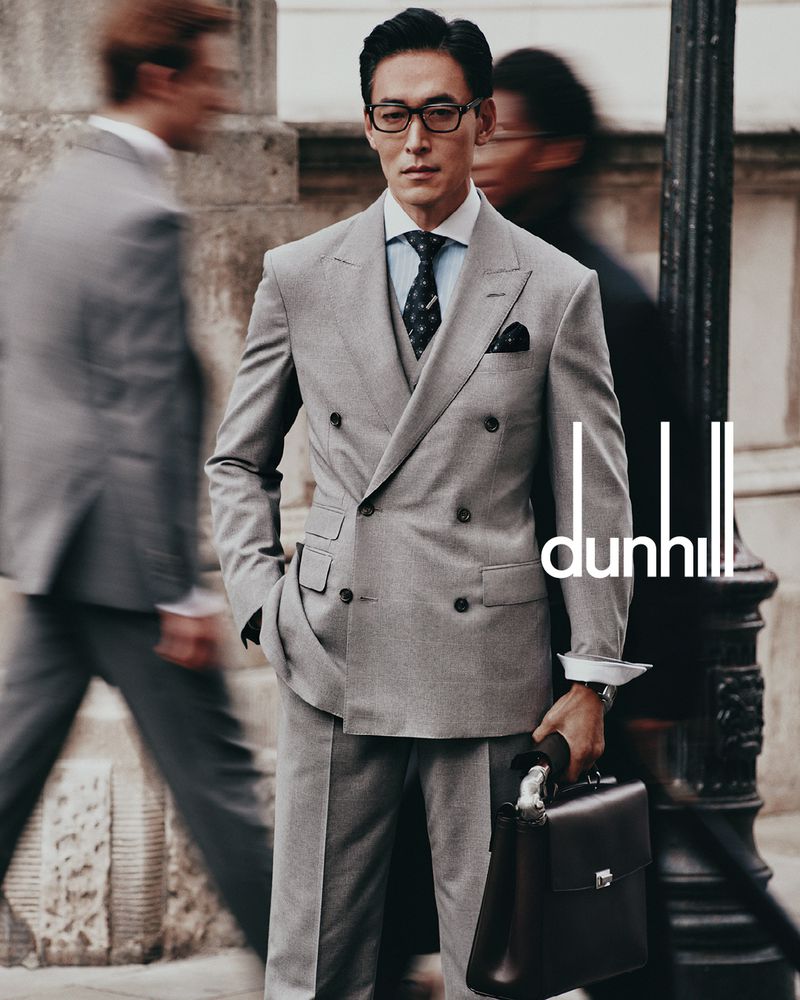 Man wearing a grey double-breasted suit and glasses holding a leather briefcase on a city street with blurred passersby.