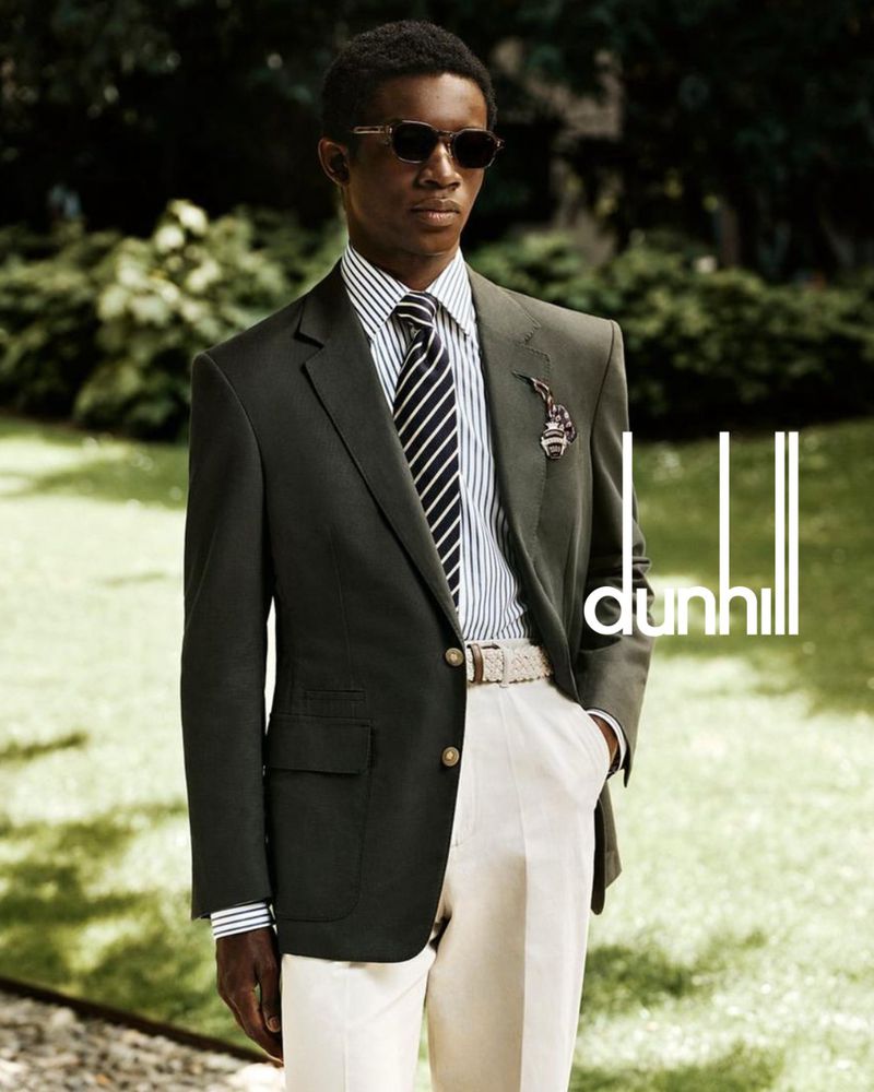 Dark-skinned man in olive blazer, striped shirt, and sunglasses stands outside wearing tailored pants and belt, Dunhill logo visible.