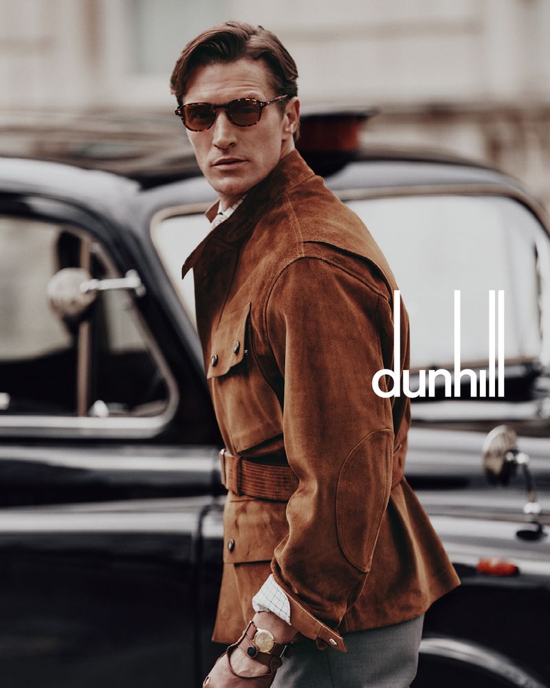 A man wearing a brown jacket, sunglasses, and a wristwatch stands next to a classic black car, with the Dunhill logo in the foreground.
