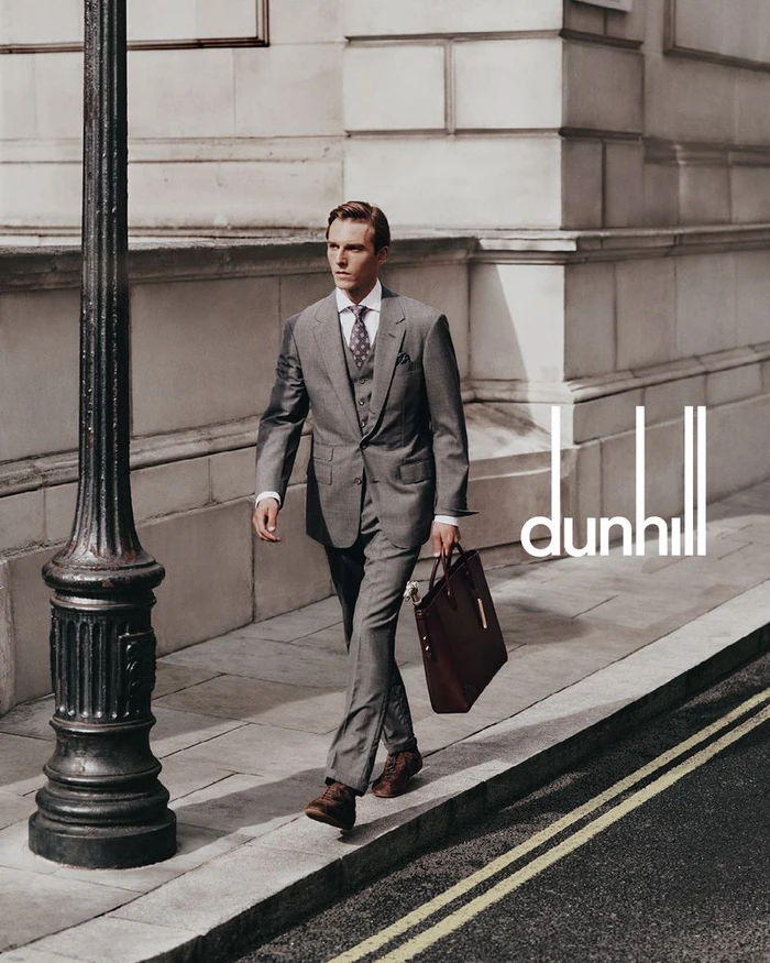 A man in a grey suit walking down a city sidewalk carrying a brown leather briefcase near a black lamppost.