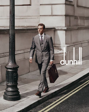 A man in a grey suit walking down a city sidewalk carrying a brown leather briefcase near a black lamppost.