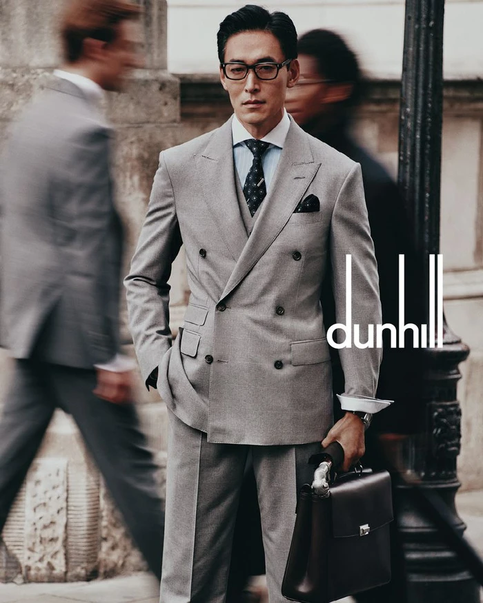 Man wearing a grey double-breasted suit and glasses holding a leather briefcase on a city street with blurred passersby.