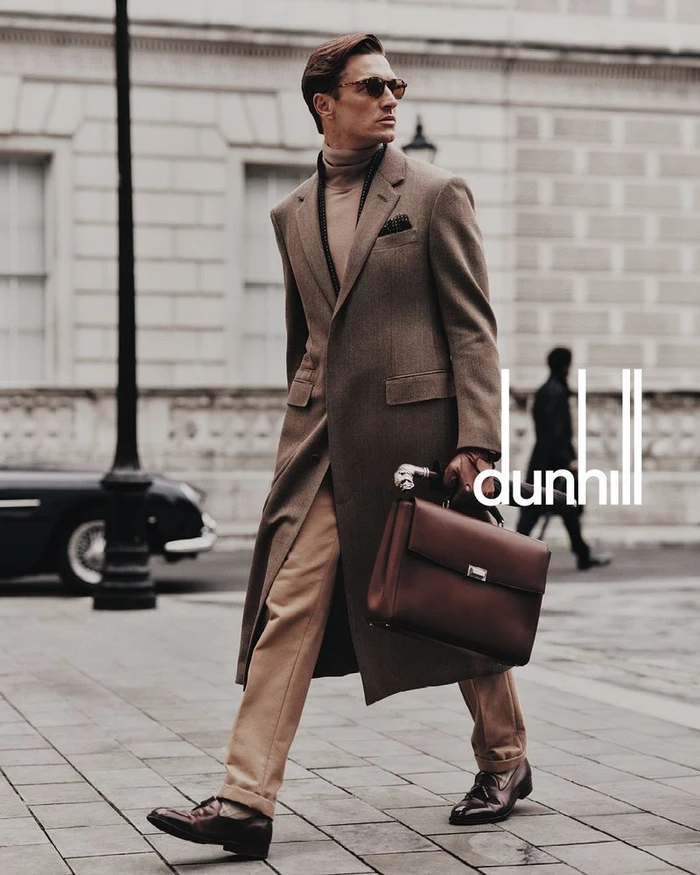 Man wearing a brown overcoat and turtleneck walking on a stone street holding a brown leather briefcase for Dunhill