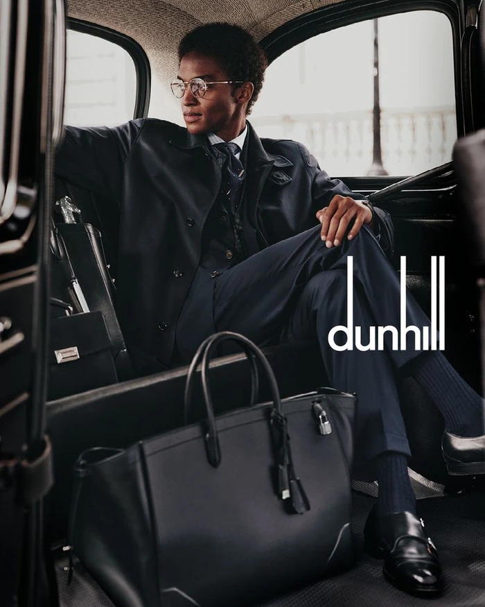 Man wearing dark suit and glasses seated inside a classic car with a black leather tote bag on the floor.