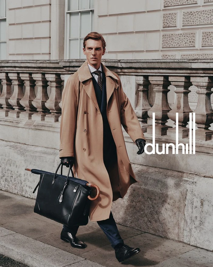 Model in a camel trench coat walking on a London street carrying a black leather holdall bag and umbrella.