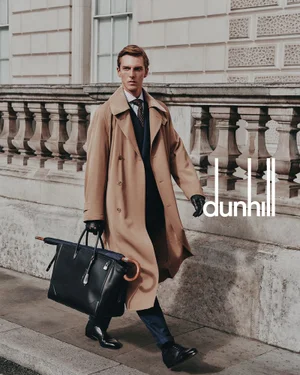 Model in a camel trench coat walking on a London street carrying a black leather holdall bag and umbrella.