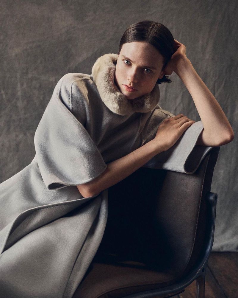 Elegant fashion portrait of a young adult female model sitting in a chair, wearing a fur-collared grey coat in a photo studio setting.