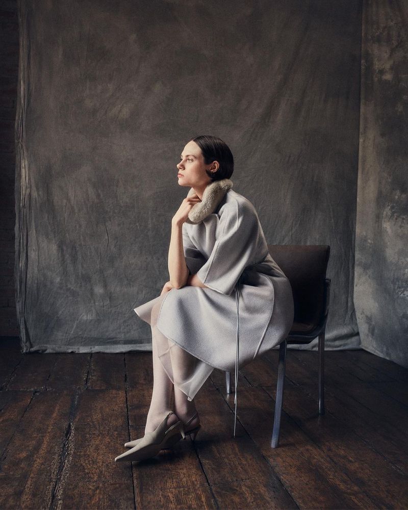 Adult woman in elegant dress and high heels sits on a chair in a photo studio against a gray backdrop, captured in soft, diffused lighting.