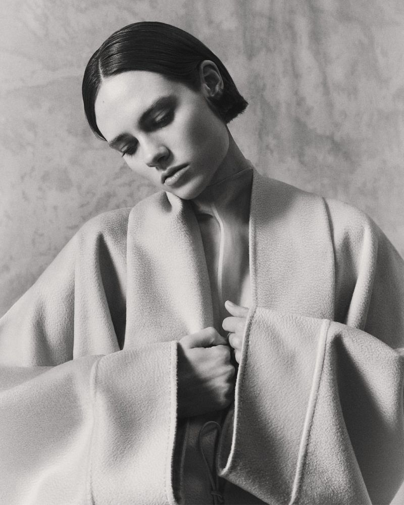 Black and white portrait of a young woman with short hair wearing a neutral coat, looking down with a calm expression in a studio setting.