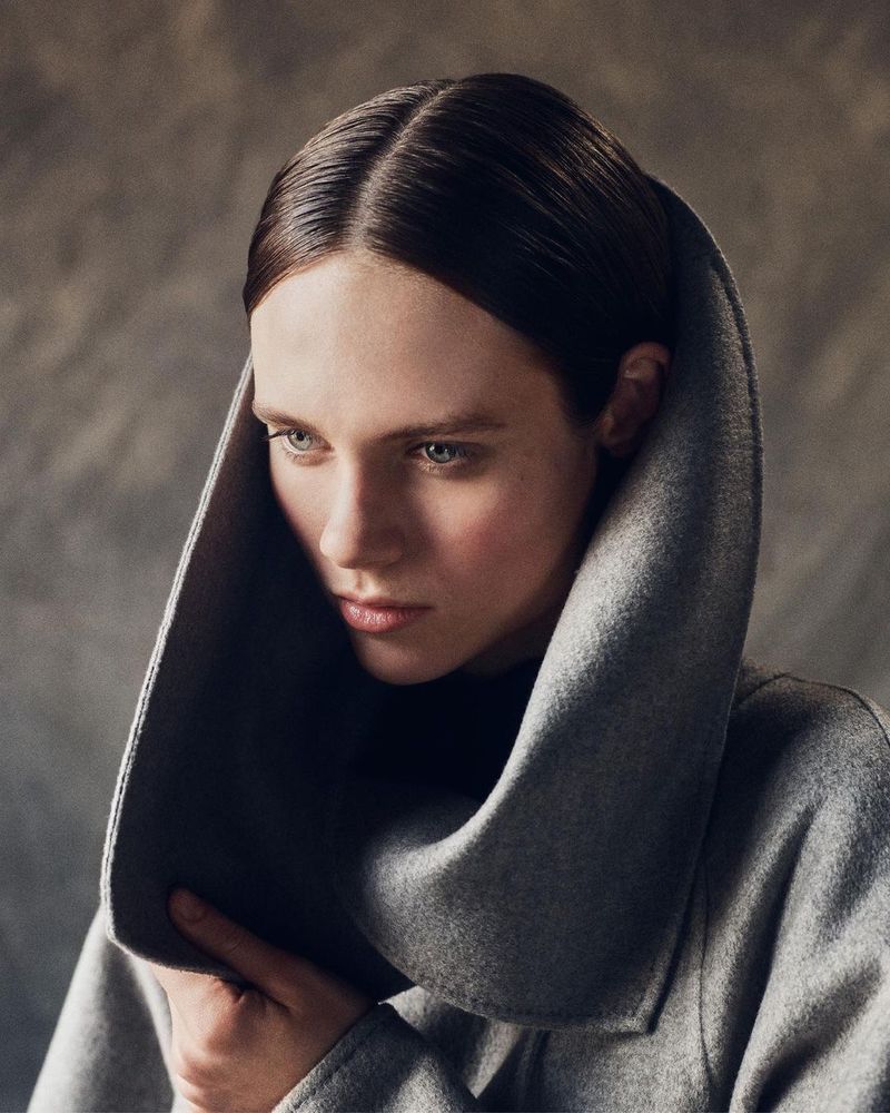 Close-up portrait of a young woman wearing a stylish gray wool coat with a large collar against a muted gray backdrop in soft lighting.