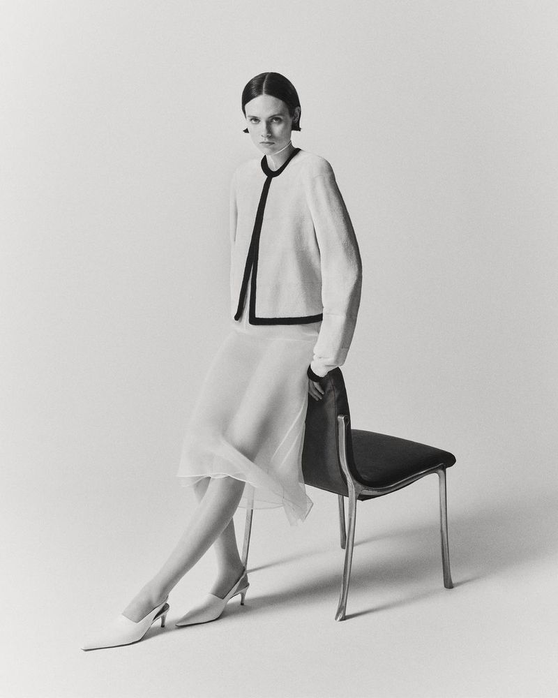 Model seated on a black chair wearing a light-coloured jacket and white skirt against a plain seamless background.