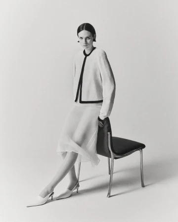 Model seated on a black chair wearing a light-coloured jacket and white skirt against a plain seamless background.