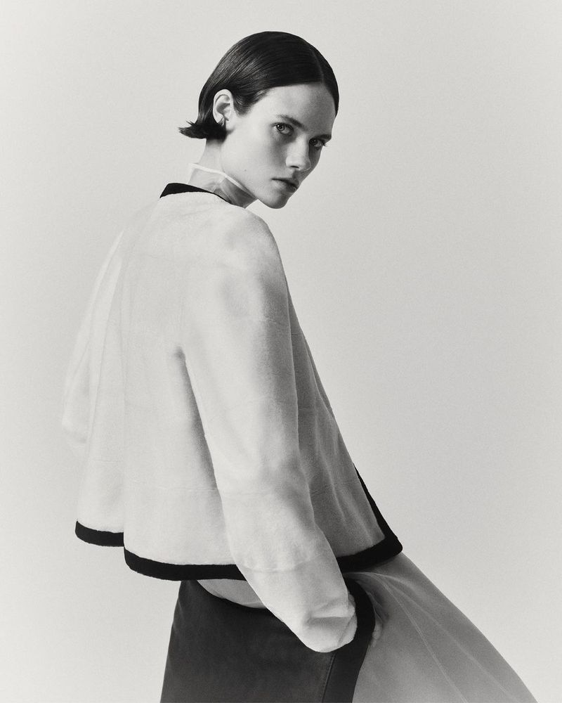 A woman with short dark hair in a light-colored top with black trim, looking over her shoulder against a neutral studio backdrop.