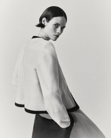 A woman with short dark hair in a light-colored top with black trim, looking over her shoulder against a neutral studio backdrop.