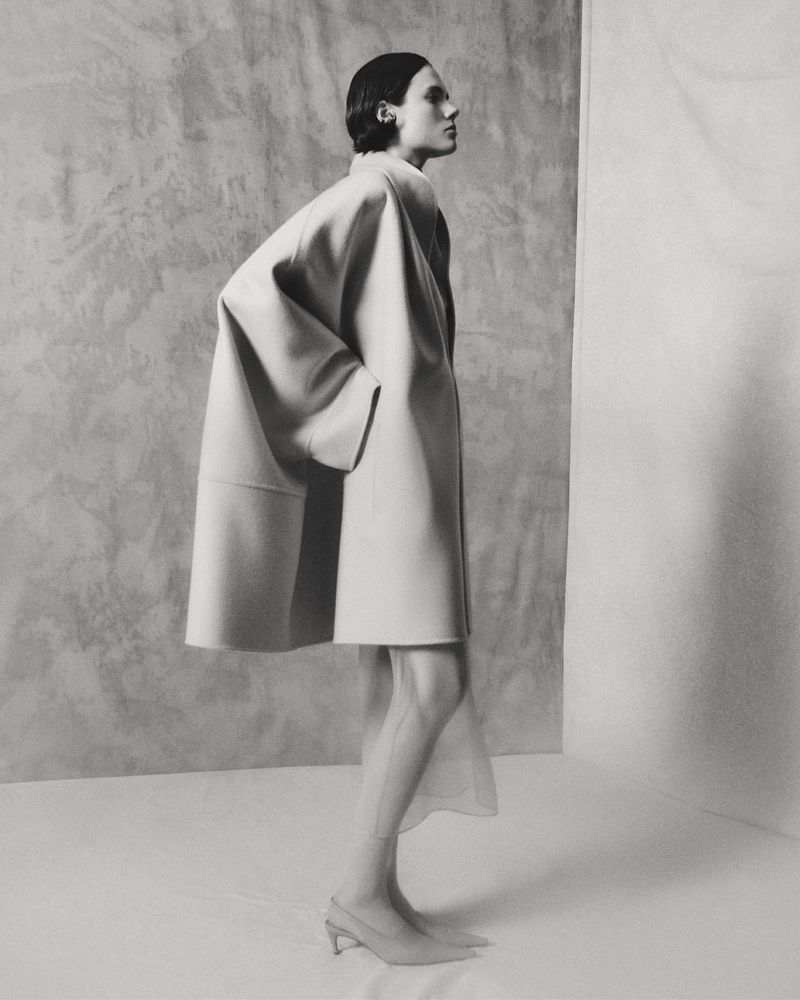 Model wearing a structured light-colored coat standing against a textured studio wall background