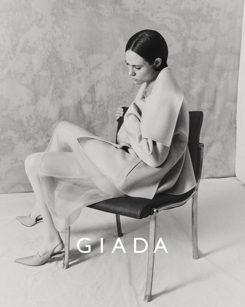 Elegant woman in sheer skirt and draped jacket sitting on a chair in a photo studio, gazing down in this monochromatic fashion shot.