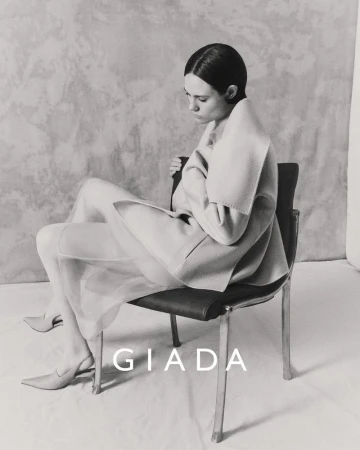 Elegant woman in sheer skirt and draped jacket sitting on a chair in a photo studio, gazing down in this monochromatic fashion shot.