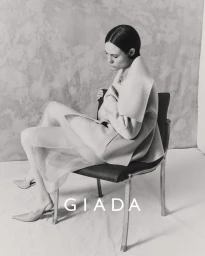 Model wearing a light-coloured coat and sheer skirt seated on a dark chair against a textured backdrop for Giada.