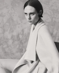Model wearing a light-coloured coat against a textured neutral background in a black-and-white portrait.