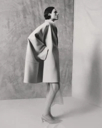 Model wearing a structured light-colored coat standing against a textured studio wall background