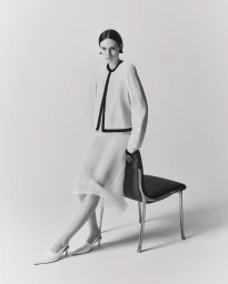 Model seated on a black chair wearing a light-coloured jacket and white skirt against a plain seamless background.