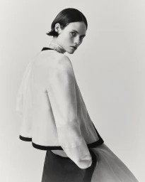 A female model wearing a cream jacket with dark trim posing against a seamless white background in a studio setting.