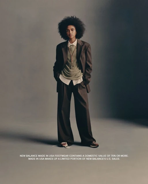 A woman with afro-textured hair poses in a brown suit and New Balance sneakers against a solid grey backdrop in a studio shot.